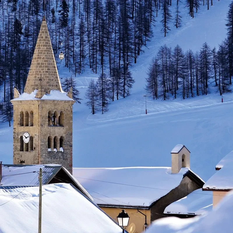Centre du village Église avec un clocher, entourée de montagnes enneigées et de maisons en bois.